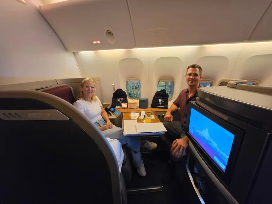 Timo und Janine sitzen sich in der Qatar Airways Kabine an einem Tisch gegenüber und lächeln.