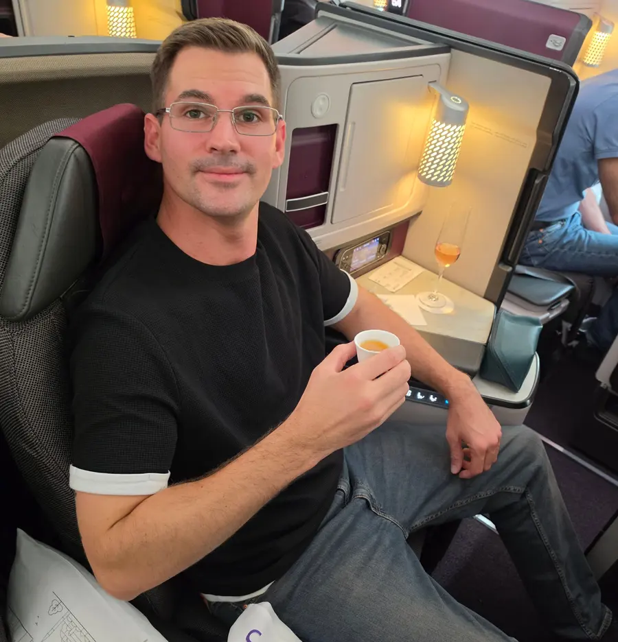 Timo sitzt im dunklen Business Sitz von Qatar Airways und hält eine kleine Tasse.