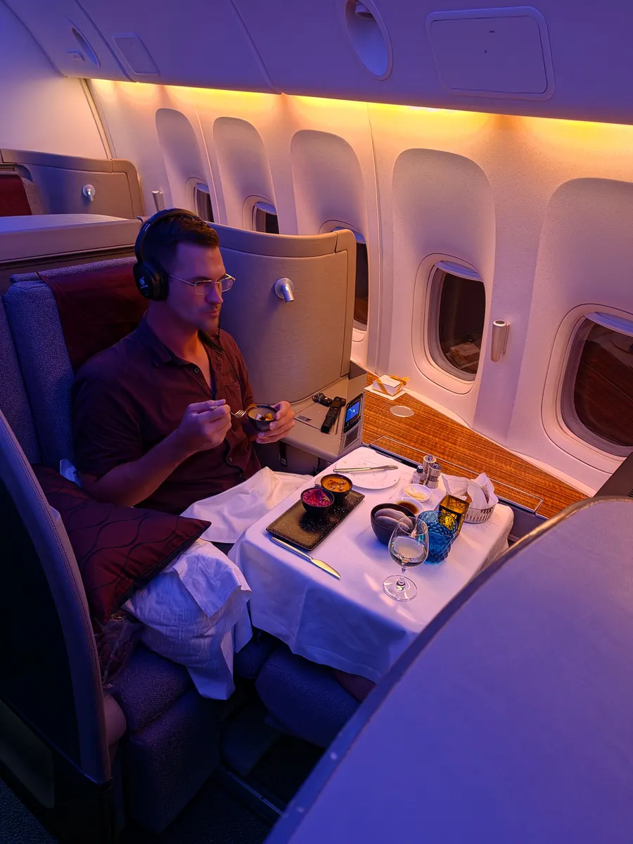 Timo sitzt in seiner Kabine im Flugzeug, trägt Kopfhörer und isst von einem gedeckten Tisch.